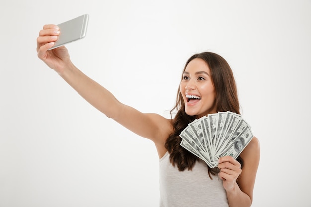 Foto della donna ricca fortunata che fa selfie sul telefono cellulare d'argento mentre tenendo un sacco di banconote in dollari dei soldi, isolata sopra la parete bianca