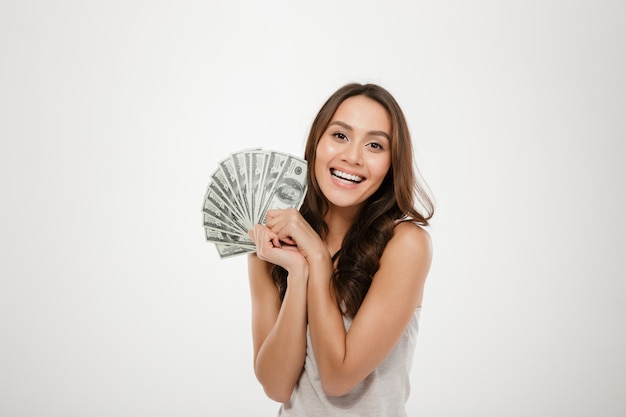 Foto della donna sorridente fortunata con capelli lunghi che vincono un sacco di banconote in dollari dei soldi, essendo ricca e felice sopra la parete bianca
