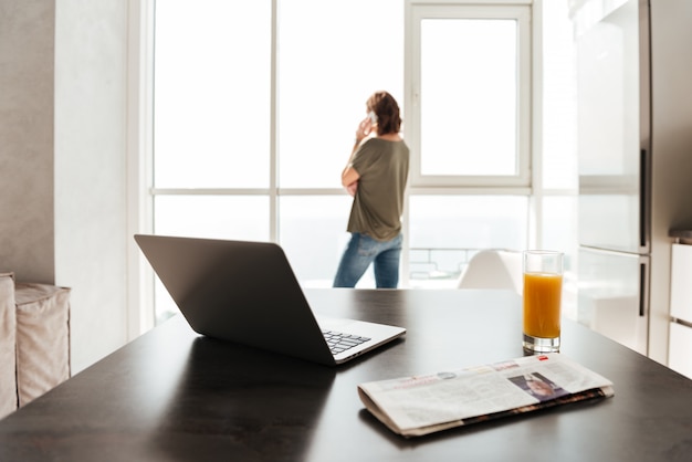 Foto della tavola con il computer portatile, il succo, il giornale e la donna vicino alla finestra