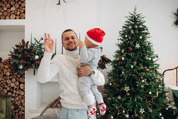 Foto della vita di un papà felice che saluta e tiene in mano un bambino vicino all'albero di Natale