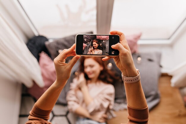 Foto delle mani che tengono il telefono durante il servizio fotografico. Ragazza dello zenzero in posa per la sua amica a casa.