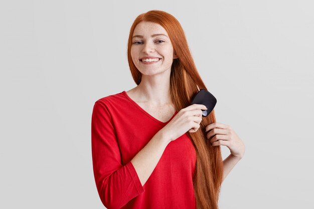 Foto di allegra sorridente giovane zenzero lentigginosa pettina i suoi lunghi capelli rossi, felice di preparare la data con il fidanzato, isolato su muro bianco. La donna si prende cura dei suoi capelli. Concetto di bellezza