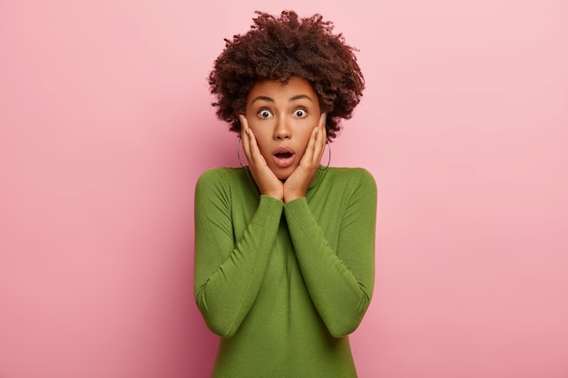 Foto di angosciata in preda al panico donna tocca le guance, guarda anxiouly, indossa poloneck verde, guarda nervosamente alla telecamera, isolato su sfondo rosa