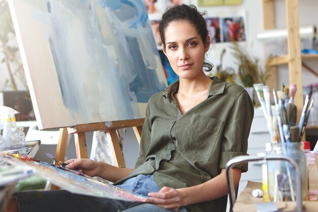 Foto di attraente giovane femmina caucasica professionale in abiti casual tenendo la tavolozza e il coltello da pittura lavorando su pittura ad olio, mescolando i colori, avendo ispirato espressione sul suo viso