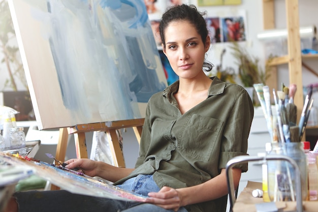 Foto di attraente giovane femmina caucasica professionale in abiti casual tenendo la tavolozza e il coltello da pittura lavorando su pittura ad olio, mescolando i colori, avendo ispirato espressione sul suo viso