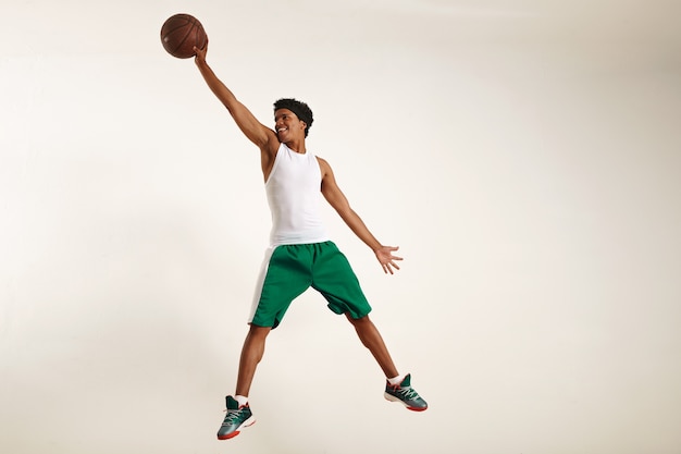 Foto di azione di un giovane atleta nero felice che indossa una camicia bianca e pantaloncini verdi che salta in alto per afferrare un pallone da basket vintage su bianco