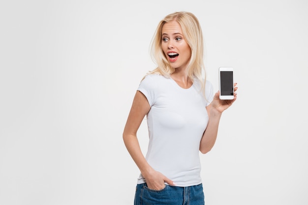 Foto di bella donna bionda con la bocca aperta e la mano in tasca, mostrando lo schermo dello smartphone in bianco