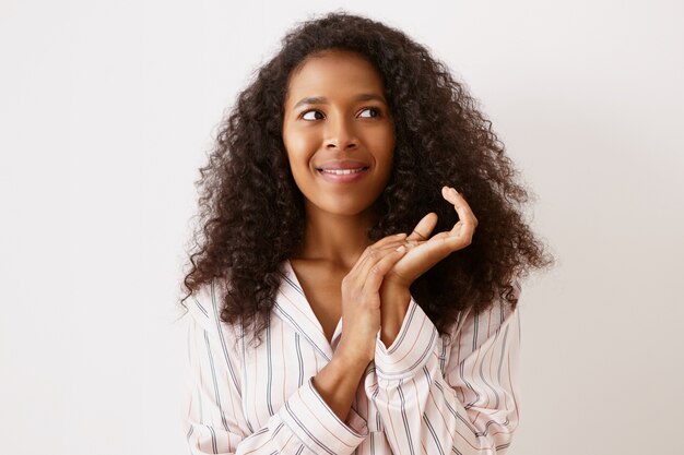 Foto di bella splendida giovane femmina dalla pelle scura con lunghi capelli voluminosi che si sente eccitata, piena di grandi idee interessanti, si strofina le mani e guarda di traverso, in posa sul muro del copyspace