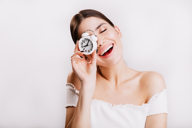 Foto di bruna sorridente senza trucco in posa con l'orologio sul muro bianco.