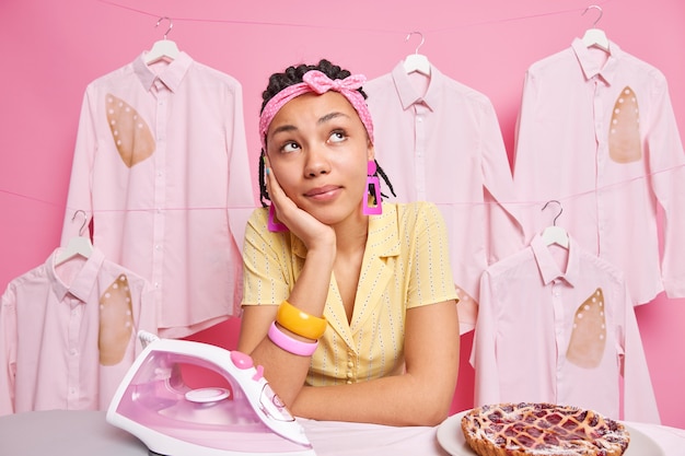 Foto di casalinga che sogna ad occhi aperti su qualcosa di impegnato a fare i lavori domestici si prende una pausa dopo aver stirato una deliziosa torta al forno vestita con abiti domestici si appoggia a bordo sul muro rosa