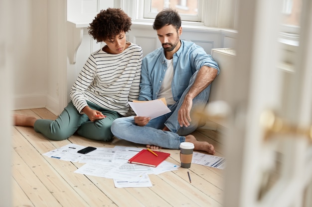 Foto di colleghi di razza mista studiano conti finanziari, indossano abiti comodi, posano sul pavimento, discutono di lavoro e collaborazione futuri, circondati da documenti, caffè e cellulare. Concetto di lavoro