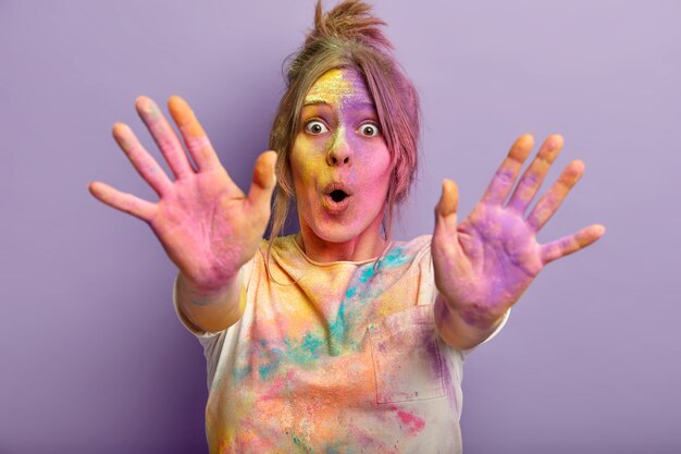 Foto di donna divertente sorpresa con viso colorato, palme e vestiti, celebra il festival di Holi, gioca con i colori, allunga le mani, isolato su un muro viola utilizza coloranti in polvere