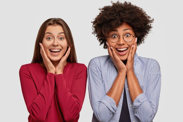 Foto di due donne interrazziali gioiose e stupite che tengono entrambe le mani sulle guance