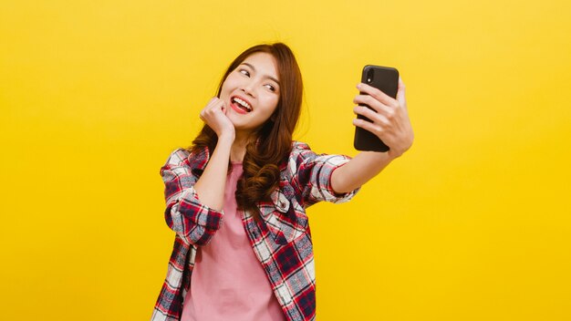 Foto di fabbricazione femminile asiatica adorabile sorridente del selfie sullo smartphone con l'espressione positiva in abbigliamento casual e sull'esame della macchina fotografica sopra la parete gialla. La donna felice adorabile felice si rallegra del successo.