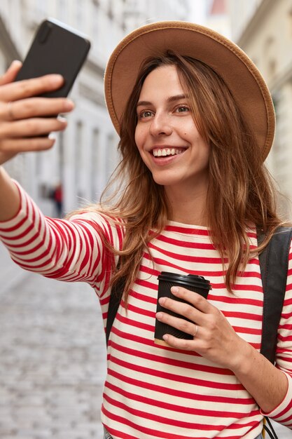 Foto di felice donna europea posa per fare selfie, ama passeggiare in centro, ricrea all'aperto