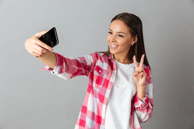 Foto di gioiosa giovane donna in camicia a scacchi, prendendo selfie dai suoi telefoni