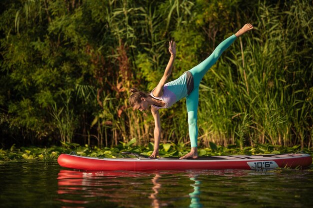 Foto di giovane donna che fa mano stare su stand up paddle board. Indossa un leggings e un top. Giornata di sole, lago blu e alberi verdi sulla riva.