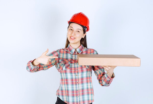 Foto di giovane donna con casco rosso che tiene in mano una scatola di cartone e indica.