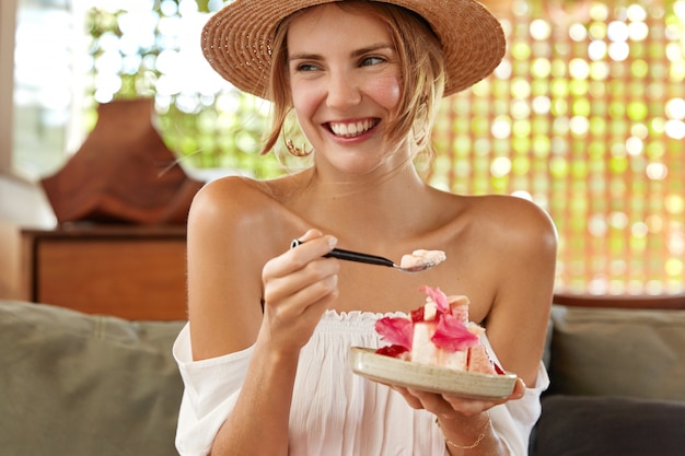 Foto di giovane donna felice con un ampio sorriso, mangia un gustoso dessert alla caffetteria, si gode la festa estiva con un amico, ascolta la storia divertente e interessante dell'interlocutore. Persone, riposo e concetto di mangiare