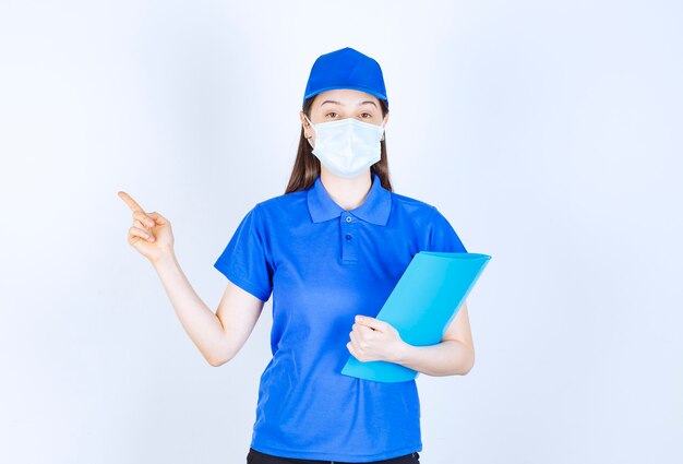 Foto di giovane donna in maschera medica che tiene cartella e punta il dito da parte.