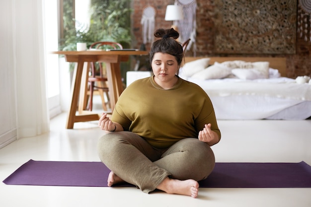 Foto di giovane donna paffuta calma tranquilla seduta a piedi nudi sulla stuoia di yoga a casa, facendo il gesto di mudra, meditando con gli occhi chiusi. Equilibrio, meditazione, armonia, zen e concetto di benessere