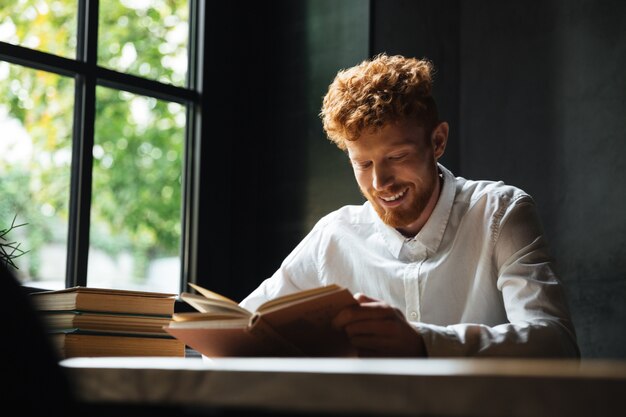 Foto di giovane uomo barbuto sorridente rossa in libro di lettura camicia bianca