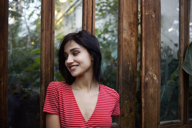 Foto di moda giovane bruna signora europea che indossa un elegante abito rosso con strisce bianche in posa all'aperto alla porta di legno con superficie a specchio, distoglie lo sguardo con positivo sorriso felice