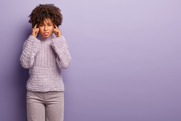 Foto di scontento donna dalla pelle scura tiene le mani sulle tempie, soffre di emicrania, sentimenti dolorosi, indossa maglione e pantaloni lavorati a maglia, cerca di concentrarsi su qualcosa. Spazio libero per il testo