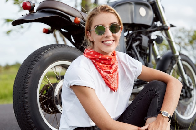 Foto di spensierata giovane motociclista professionista indossa occhiali da sole alla moda e bandana, si siede vicino a una moto nera veloce, ama la guida all'aperto, si siede sull'asfalto vicino al suo mezzo di trasporto preferito