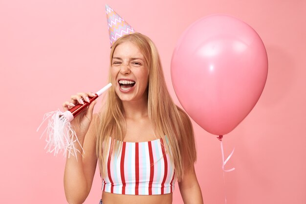 Foto di splendida ragazza adolescente carina con capelli biondi sciolti e bretelle in posa su rosa con ventilatore di partito e palloncino a elio, ridendo con la bocca spalancata