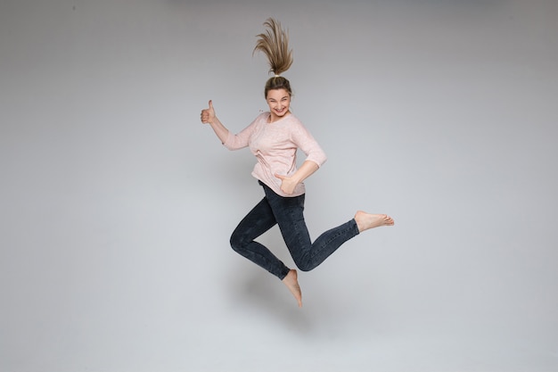 Foto di stock di allegra spensierata donna bionda che salta a mezz'aria con il pollice in alto su sfondo bianco. Donna di salto che sorride alla macchina fotografica che tiene i pollici in su.