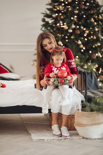 Foto di stock di amorevole madre in abito verde dando la sua piccola figlia in pigiama vestire un regalo di Natale. Sono accanto all'albero di Natale splendidamente decorato sotto la nevicata.