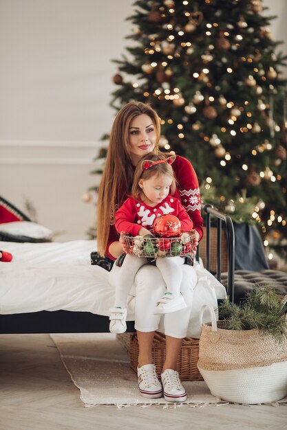 Foto di stock di amorevole madre in abito verde dando la sua piccola figlia in pigiama vestire un regalo di Natale. Sono accanto all'albero di Natale splendidamente decorato sotto la nevicata.
