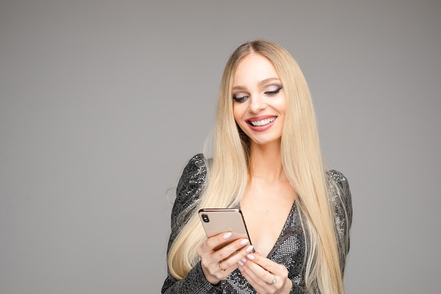 Foto di stock di splendida donna bionda con i capelli lunghi in abito da cocktail grigio navigando sul cellulare, divertendosi e sorridendo.