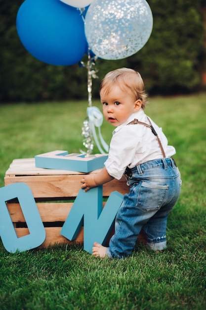 Foto di stock di un adorabile ragazzino in jeans, maglietta e bretelle che gioca con decorazioni di compleanno sul prato nel cortile di casa. Giornata estiva fuori. Concetto di compleanno. Mongolfiere e lettere in legno.