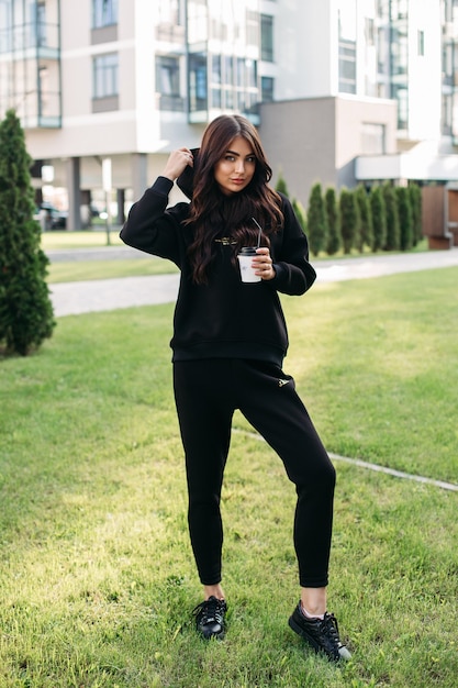 Foto di stock di una bella ragazza sportiva in maglione nero e jogging che indossa un cappuccio mentre si tiene una tazza di caffè per andare. Ragazza alla moda in abiti sportivi neri e scarpe da ginnastica in piedi sul prato verde.