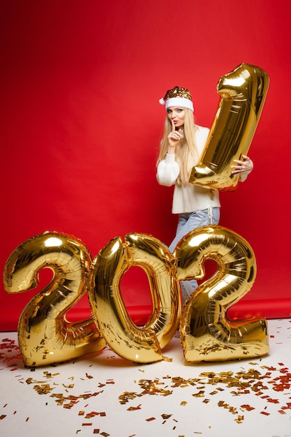 Foto di stock di una ragazza bionda con un cappello da Babbo Natale e un maglione che ruba la cifra gonfiabile numero uno e scappa di fronte alle cifre del nuovo anno. Sfondo rosso.