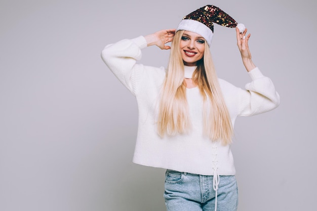 Foto di stock ritratto di bella donna bionda in maglione bianco che indossa brillante cappello di Babbo Natale scintillante in posa su sfondo bianco. Copia spazio.