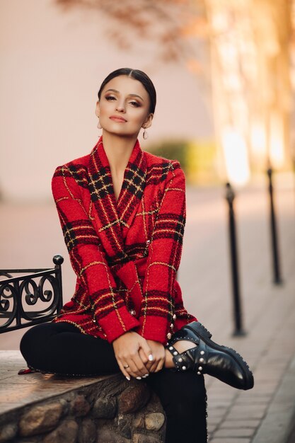 Foto di stock ritratto di giovane ragazza attraente in luminoso trench a quadri, pantaloni neri e stivali alla moda seduto su una panchina nel parco. Guardando la fotocamera con aria sognante.