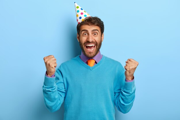 Foto di studio di ragazzo felicissimo con cappello di compleanno in posa in maglione blu