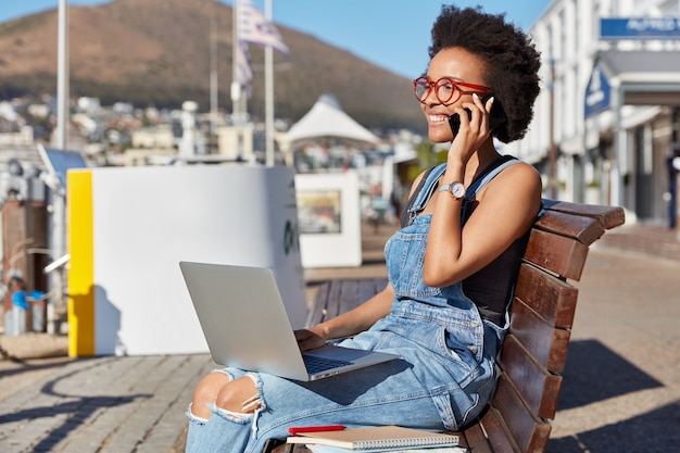 Foto di un adolescente afroamericano felice e sorridente chiama qualcuno tramite il cellulare, tiene il computer portatile sulle ginocchia, si siede a una panchina all'aperto usa gadget per studiare online, bloggs. Moda, lifestyle, tecnologia