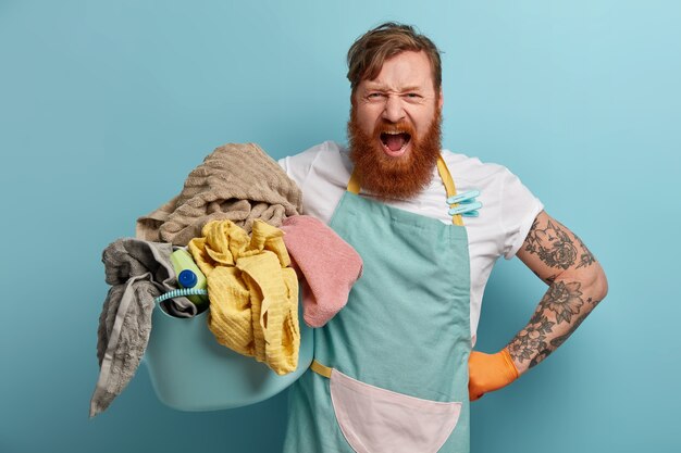 Foto di un uomo barbuto infastidito impegnato con i lavori domestici, tiene un cesto pieno di bucato e detersivo, indossa un grembiule, grida ad alta voce, si sente infastidito, isolato sul muro blu, stanco di fare il bucato.