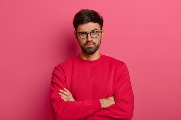 Foto di un uomo sicuro di sé con la barba, tiene le braccia incrociate sul corpo, guarda seriamente, indossa abiti casual, parla con un collega, posa al chiuso contro il muro rosa. Ragazzo fiducioso