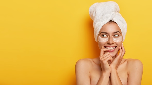 Foto di una bella giovane donna con la pelle fresca e sana, distoglie lo sguardo, ha procedure di bellezza, sta con le spalle nude contro il muro giallo, si prende cura del suo corpo, nutre la pelle e indossa le toppe per la morbidezza