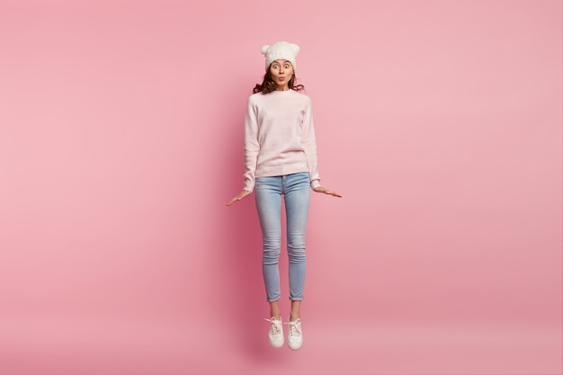 Foto di una bella ragazza che salta in aria contro lo spazio rosa, indossa cappello, maglione, jeans e scarpe da ginnastica, tiene le labbra piegate