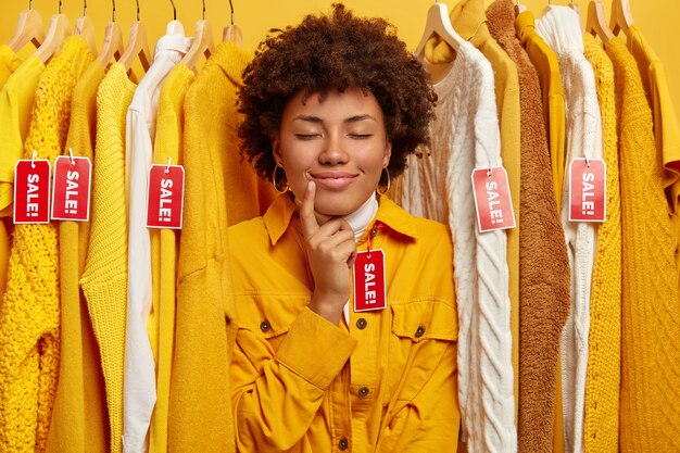 Foto di una donna adorabile con un taglio di capelli afro, prova la nuova giacca gialla nel negozio di vestiti, tiene gli occhi chiusi, si trova tra i vestiti con i cartellini rossi in vendita, cerca un vestito alla moda.