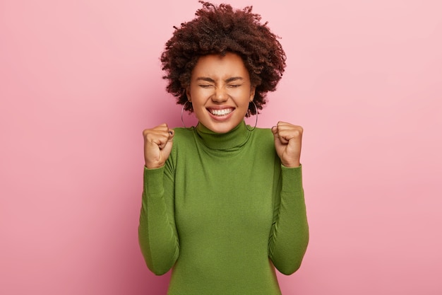 Foto di una donna afro felicissima stringe i pugni con trionfo, sorride ampiamente, si rallegra di nuovi risultati