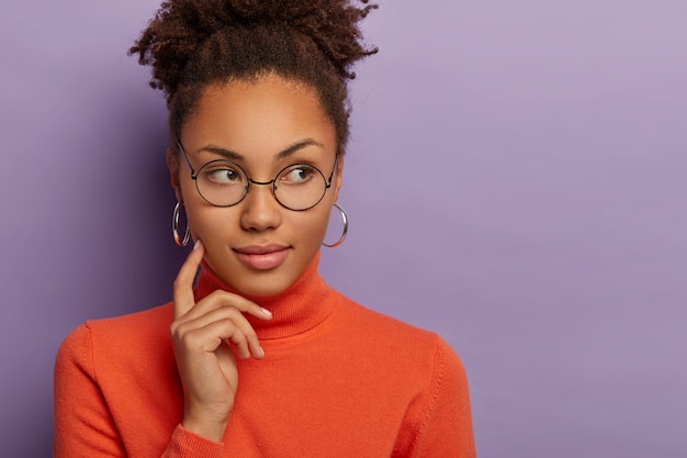 Foto di una donna afroamericana premurosa che tiene il dito indice sulla guancia, guarda da parte con espressione pensierosa, pensa a cosa fare, indossa occhiali rotondi, orecchini d'argento e poloneck, modella indoor