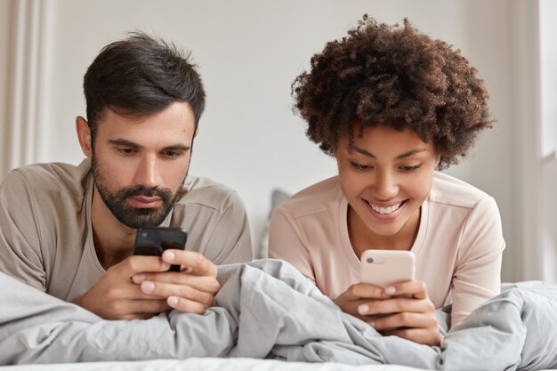 Foto di una donna e un uomo dipendenti di razze diverse, utilizzare i telefoni cellulari a letto, connessi a Internet wireless, godersi la mattina nel fine settimana, avere espressioni allegre. Concetto di moderne tecnologie