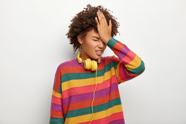 Foto di una donna etnica stressante schiaffeggia la fronte con il palmo, dimentica informazioni importanti o si rammarica di aver fatto qualcosa di sbagliato, indossa un maglione multicolore, cuffie